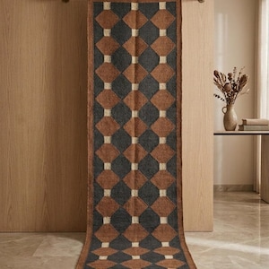 May include: A long, rectangular runner rug featuring a repeating geometric pattern of dark charcoal grey and terracotta brown diamonds, separated by small cream-coloured squares. The rug has a textured weave and short fringe at both ends.