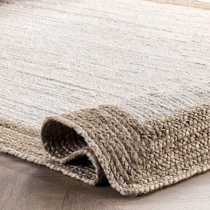 May include: A natural fiber rug with a rolled-up edge. The rug features a two-tone design, with a light beige center and a darker brown border. The texture appears woven, adding a rustic and textured element to the home decor.