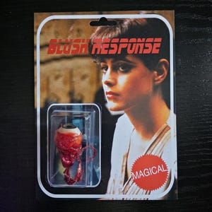 May include: A novelty item presented on a retro-style card. The card features a woman's portrait and the text "BLUSH RESPONSE." A plastic blister pack contains a realistic-looking, bloodshot eyeball with attached veins. A red sticker with the word "MAGICAL" is on the card.