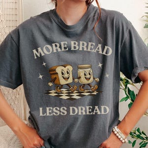 May include: A charcoal gray t-shirt with the words "MORE BREAD LESS DREAD" in a vintage font. The shirt features a cartoon illustration of a slice of bread and a jar of peanut butter walking on a checkered floor.