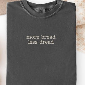 May include: Dark grey t-shirt with the text "more bread less dread" in a simple, sans-serif font. The shirt has a crew neck and a slightly faded appearance. The shirt is folded neatly on a light-colored surface.