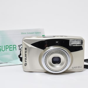 May include: A silver Fujifilm Super 120 AZ 35mm compact camera with a black accent and a built-in flash. The camera has a zoom lens and a wrist strap. A manual with the text "SUPER 12" is to the left.