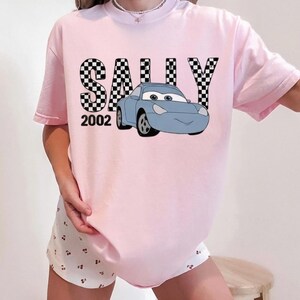 May include: A pale pink t-shirt with the name "SALLY" in a black and white chequered design, a blue cartoon car, and the number "2002". The shirt has short sleeves and a casual fit.