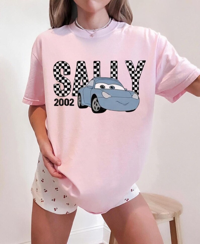 May include: A pale pink t-shirt with the name "SALLY" in a black and white chequered design, a blue cartoon car, and the number "2002". The shirt has short sleeves and a casual fit.