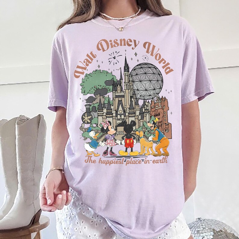 May include: A light purple t-shirt featuring a colorful graphic of Disney World characters and landmarks. The design includes Mickey and Minnie Mouse, Donald Duck, Goofy, and the iconic Cinderella Castle. Text reads "Walt Disney World" and "The happiest place on earth."