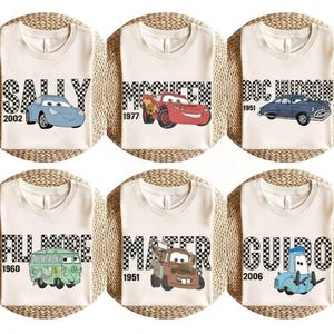 May include: Six cream-coloured t-shirts featuring cartoon car characters. Each shirt has a different car name in a black and white chequered font, with the car's image and a year. The cars include Sally, McQueen, Doc Hudson, Fillmore, Mater, and Guido.