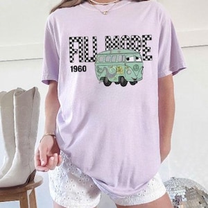 May include: A lilac t-shirt with a cartoon green van graphic and the text "FILL MODE 1960". The words "FILL MODE" are in a black and white chequered pattern. The van has a smiling face and floral details.