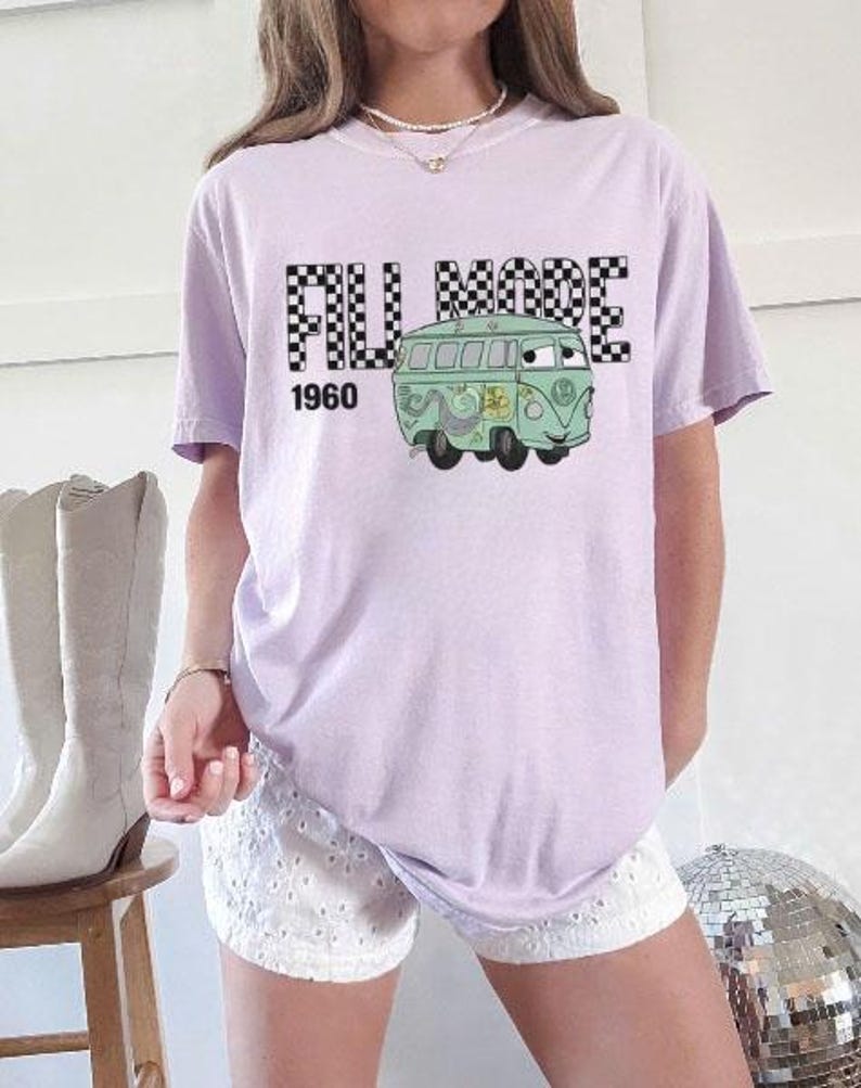 May include: A lilac t-shirt with a cartoon green van graphic and the text "FILL MODE 1960". The words "FILL MODE" are in a black and white chequered pattern. The van has a smiling face and floral details.