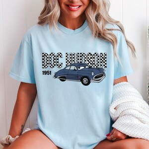 May include: Pale blue t-shirt with a cartoon illustration of a blue car and the text "DOC HUDSON 1951". The car has a smiling face and is a vintage style. The text "DOC HUDSON" is in a black and white checkered pattern. Casual wear.