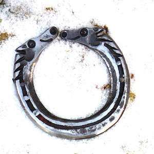 May include: A horseshoe-shaped metal sculpture featuring two horse heads facing each other. The horseshoe is silver with dark accents and is set against a snowy background. The sculpture is likely a decorative item.