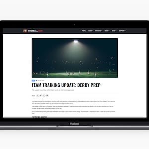 May include: A MacBook laptop shows a Football Live webpage with the headline "TEAM TRAINING UPDATE: DERBY PREP". The image on the webpage shows a football team training on a green pitch under bright stadium lights. The laptop is on a white surface.