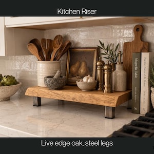 May include: A kitchen riser featuring a rectangular live edge oak wood slab supported by four short, square-profile steel legs. The natural wood grain is visible on the top surface, which holds various kitchen items like bowls and utensils.
