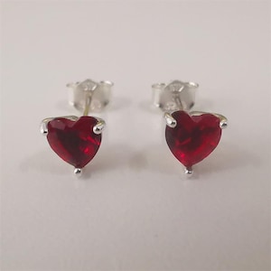 Pode incluir: Um par de brincos de tarracha com pedras vermelhas facetadas em forma de coração, montadas em garras de metal prateado. Cada brinco inclui um fecho de borboleta a condizer, exibindo um design clássico com um tom carmesim vibrante.