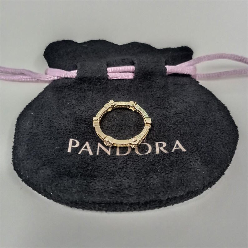 May include: A gold-colored ring with small, clear stones is displayed on a black pouch with the word "PANDORA" printed in white. The ring has a repeating design of rectangular and curved elements.