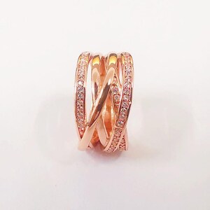 Puede incluir: Anillo de color oro rosa con un dise&ntilde;o entrelazado. El anillo presenta m&uacute;ltiples bandas que se cruzan, con una banda incrustada con peque&ntilde;as piedras preciosas transparentes. El anillo est&aacute; sobre un fondo blanco.