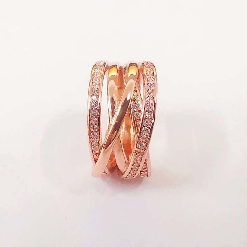 Puede incluir: Anillo de color oro rosa con un dise&ntilde;o entrelazado. El anillo presenta m&uacute;ltiples bandas que se cruzan, con una banda incrustada con peque&ntilde;as piedras preciosas transparentes. El anillo est&aacute; sobre un fondo blanco.