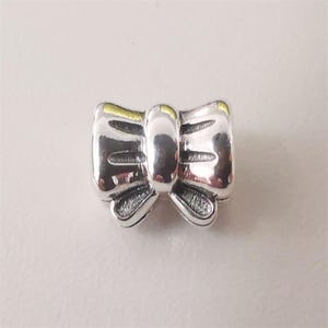 Könnte beinhalten: Ein polierter silberfarbener Metall-Charm-Bead in Form einer Schleife. Der Anhänger hat eine glatte, reflektierende Oberfläche mit einem zentralen vertikalen Band und zwei symmetrischen Schlaufen an den Seiten sowie texturierten Details an den unteren Kanten.
