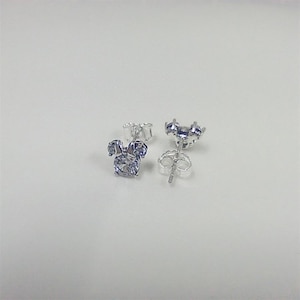 May include: A pair of silver-colored stud earrings. Each earring features a three-stone design, with a larger round stone at the center and two smaller stones above, resembling a cartoon character's head. The earrings are set against a white background.