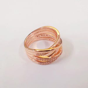 Puede incluir: Anillo de color oro rosa con un dise&ntilde;o entrelazado. El anillo presenta m&uacute;ltiples bandas que se cruzan, con una fila de peque&ntilde;as piedras brillantes incrustadas en una de las bandas. El anillo est&aacute; sobre un fondo blanco.