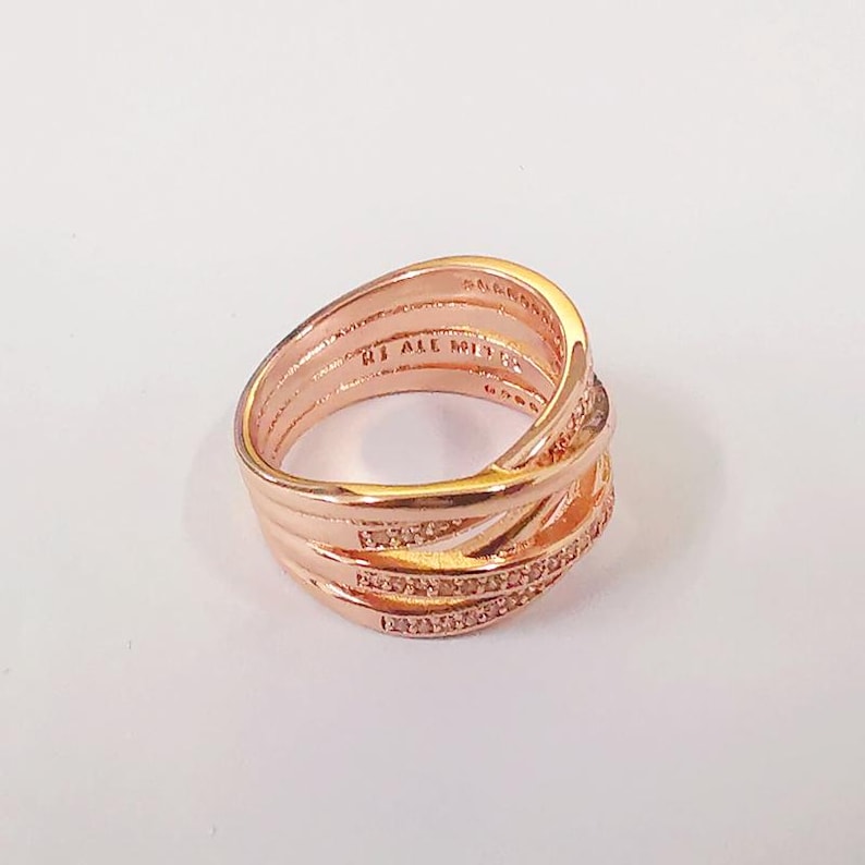 Puede incluir: Anillo de color oro rosa con un dise&ntilde;o entrelazado. El anillo presenta m&uacute;ltiples bandas que se cruzan, con una fila de peque&ntilde;as piedras brillantes incrustadas en una de las bandas. El anillo est&aacute; sobre un fondo blanco.
