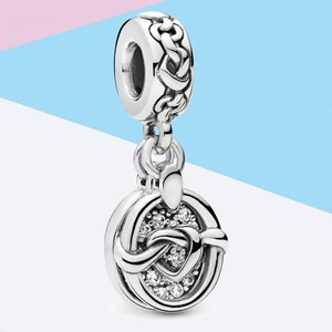 May include: A silver charm with a Celtic knot design. The charm features a braided top, a circular pendant with a knot design, and small, clear gemstones. The charm is suspended from a ring and is suitable for a bracelet or necklace.