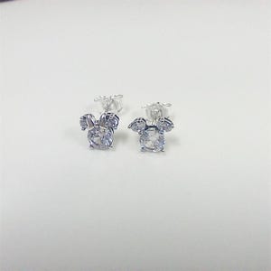 May include: A pair of silver-toned stud earrings. Each earring features a large, round, clear gemstone at the center, surrounded by smaller, clear gemstones arranged in a bow shape. The earrings are set against a white background.
