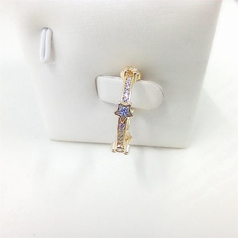 May include: A gold-colored ring with a star-shaped blue gemstone in the center. The ring is adorned with small, clear gemstones. The ring is displayed on a white surface.