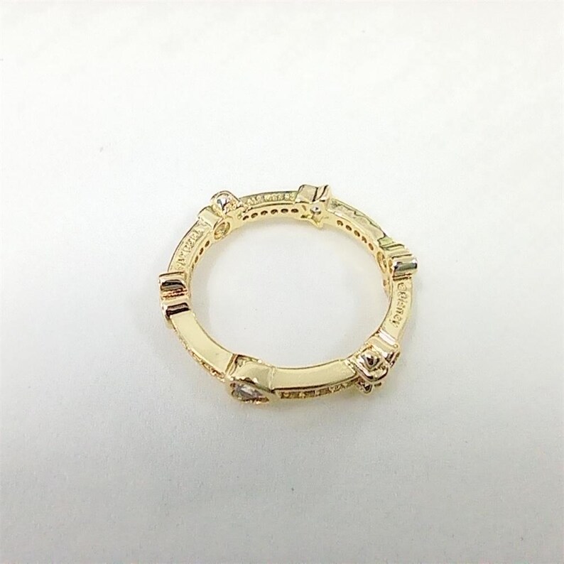 May include: A gold-colored ring with a repeating geometric design. The ring features small, clear gemstones and is set against a white background. The word "Elegance" is visible on the ring.