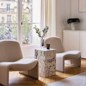 Può includere: Scena di un soggiorno moderno con due poltrone beige e un tavolino in marmo. Una lampada in bianco e nero su un mobile bianco. Un vaso con verde e un bicchiere sul tavolino. Grande finestra.