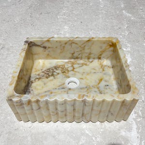 Peut inclure: Évier rectangulaire en marbre avec bordure festonnée. L'évier présente un motif marbré crème et marron et un trou d'évacuation central. Un élément décoratif pour une salle de bain.
