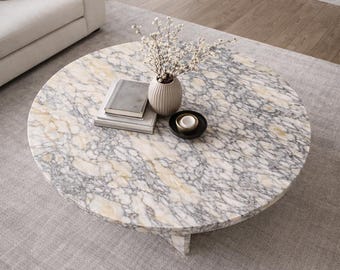 May include: A round marble coffee table with a gray and cream pattern. The table is topped with a stack of books, a small vase with white flowers, and a black dish. The table sits on a gray rug.