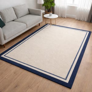 May include: A rectangular rug with a cream centre and a navy blue border. The border has a double stripe design in navy and light blue. The rug is on a wooden floor in a living room setting, near a sofa.