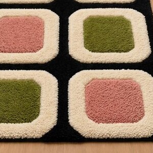 以下が含まれることがあります： 丸みを帯びた正方形が繰り返される幾何学模様のプラッシュラグ。ピンクとオリーブグリーンのテクスチャー加工された正方形が交互に配置され、それぞれが黒い背景の上にクリーム色の糸の厚い縁取りで囲まれています。