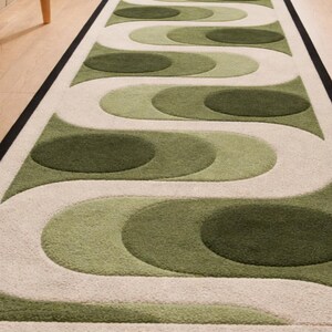 Puede incluir: Una alfombra larga y rectangular con un patr&oacute;n repetitivo de formas verdes y blanquecinas. La alfombra tiene un borde negro y est&aacute; colocada sobre un suelo de madera. El dise&ntilde;o presenta c&iacute;rculos superpuestos y l&iacute;neas curvas.