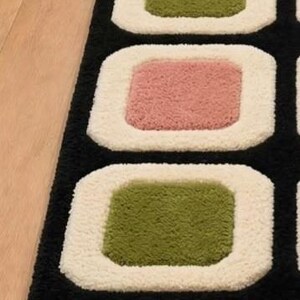 以下が含まれることがあります： 黒い背景に丸みを帯びた四角形が繰り返されるデザインの長いランナーラグ。各四角形は、厚みのあるクリーム色の縁取りが特徴で、中央部分は落ち着いたオリーブグリーンとダスティローズピンクが交互に配置されています。