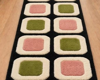 Puede incluir: Una alfombra de pasillo larga con fondo negro y dos filas paralelas de formas cuadradas repetitivas. Cada cuadrado tiene un borde grueso y texturizado de color crema que rodea un bloque central sólido en tonos alternos de verde oliva y rosa.