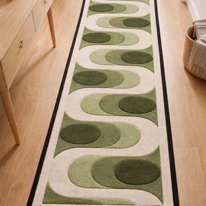Puede incluir: Una alfombra rectangular con dise&ntilde;o retro. La alfombra presenta un patr&oacute;n repetitivo de formas verdes y blanquecinas con un borde negro. La alfombra est&aacute; colocada sobre un suelo de madera.