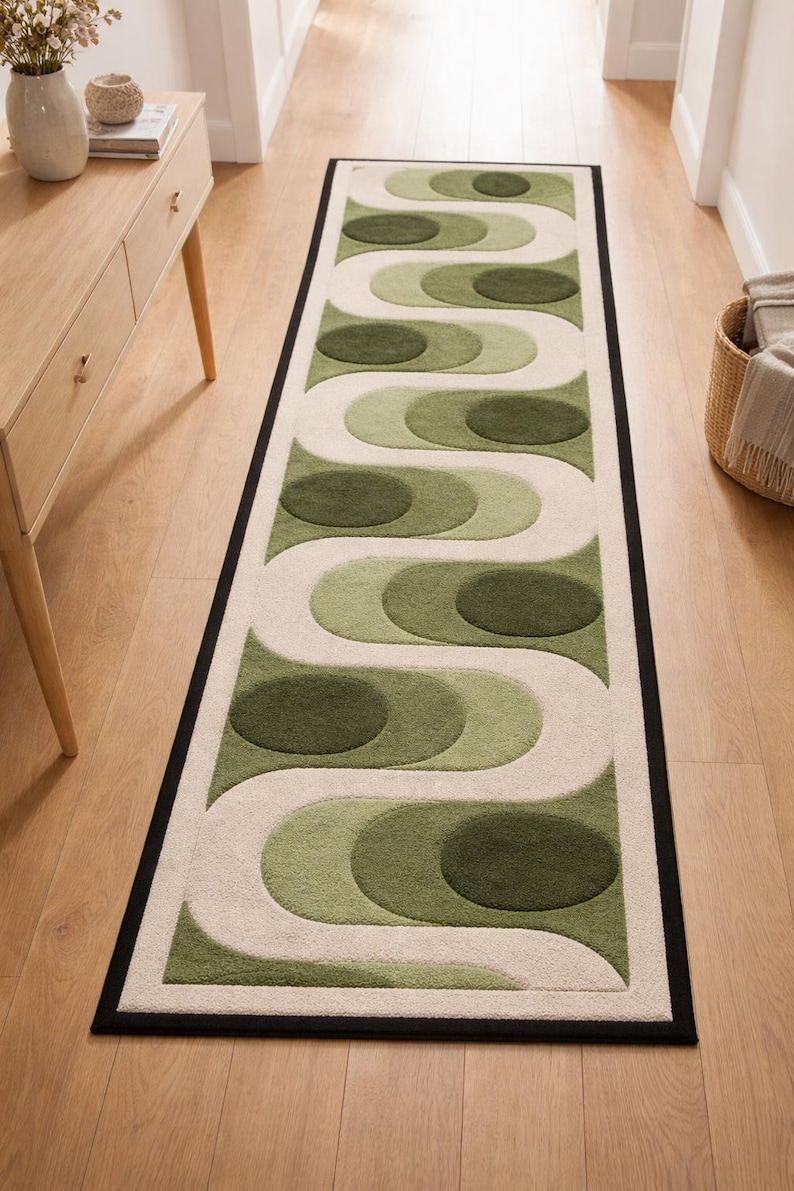 Puede incluir: Una alfombra rectangular con dise&ntilde;o retro. La alfombra presenta un patr&oacute;n repetitivo de formas verdes y blanquecinas con un borde negro. La alfombra est&aacute; colocada sobre un suelo de madera.
