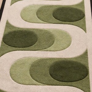 Puede incluir: Una alfombra rectangular con un dise&ntilde;o retro. La alfombra presenta un patr&oacute;n repetitivo de formas verdes y crema, incluyendo c&iacute;rculos y l&iacute;neas curvas. La alfombra tiene un borde negro.