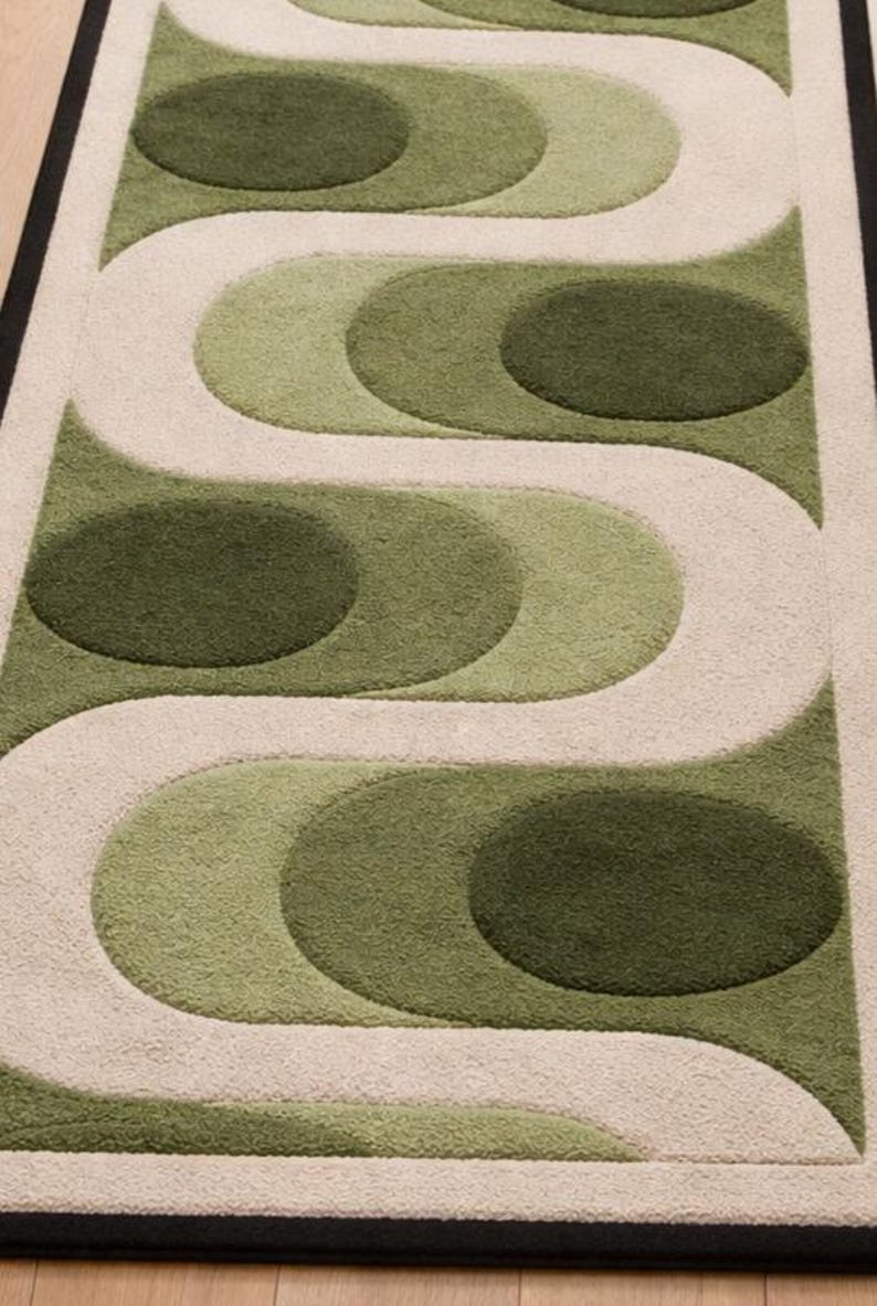 Puede incluir: Una alfombra rectangular con un dise&ntilde;o retro. La alfombra presenta un patr&oacute;n repetitivo de formas verdes y crema, incluyendo c&iacute;rculos y l&iacute;neas curvas. La alfombra tiene un borde negro.