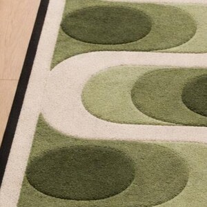 Puede incluir: Una alfombra estampada en verde y blanco roto con un borde negro. La alfombra presenta un dise&ntilde;o repetitivo de l&iacute;neas curvas y formas ovaladas en diferentes tonos de verde. La alfombra est&aacute; sobre un suelo de madera.