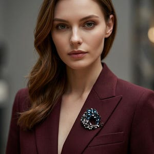 Könnte beinhalten: Ein bordeauxfarbener Blazer mit einer dekorativen Brosche. Die Brosche hat ein dunkelblaues und weißes Design. Die Frau auf dem Bild hat lange braune Haare und schaut direkt in die Kamera.