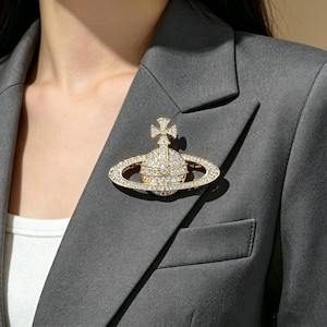Könnte beinhalten: Goldfarbene Brosche in Form eines Planeten mit Kreuz, besetzt mit klaren Kristallen. Die Brosche ist an einem grauen Blazer befestigt, was einen auffälligen Kontrast erzeugt. Das Design ist aufwendig und detailliert.