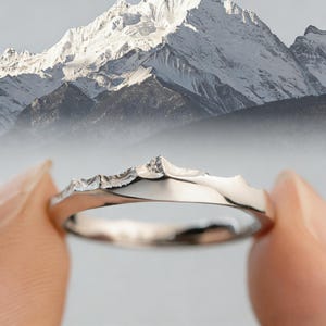 Könnte beinhalten: Ein silberner Ring mit einem Gebirgsmotiv. Der Ring hat ein glattes, poliertes Band, während die Berggipfel eine strukturierte, raue Oberfläche aufweisen. Der Hintergrund zeigt ein verschwommenes Bild von schneebedeckten Bergen.