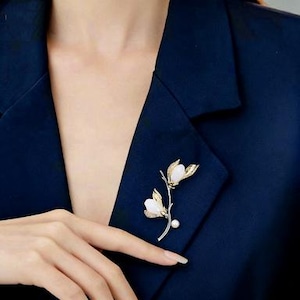 Könnte beinhalten: Goldfarbene Blumenbrosche mit weißen Blütenblättern und einer Perle, an einem marineblauen Blazer befestigt. Die Brosche hat zarte Blattdetails und wird am Revers getragen.