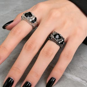 Könnte beinhalten: Zwei Ringe mit schwarzen Edelsteinen. Einer ist silberfarben mit strukturiertem Design, der andere dunkelgrau mit ähnlichem Muster. Die Ringe werden an Fingern getragen und präsentieren den Schmuck.