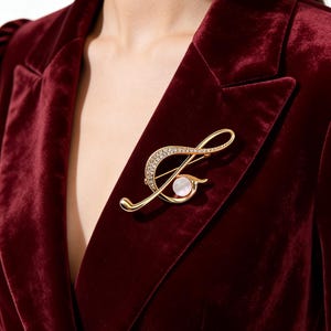 Könnte beinhalten: Goldfarbene Brosche in Form einer Musiknote, verziert mit klaren Kristallen und einem perlenartigen Akzent. Die Brosche ist an einem bordeauxfarbenen Samtblazer befestigt und bildet einen auffälligen Kontrast. Das Design ist elegant und für formelle Anlässe geeignet.