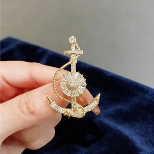 Könnte beinhalten: Goldfarbene Anstecknadel in Ankerform mit funkelnden, klaren Kristallen. Das Ankerdesign weist ein Seildetail und einen zentralen Sonnenstrahl aus Kristallen auf. Ein stilvolles Accessoire im maritimen Stil.