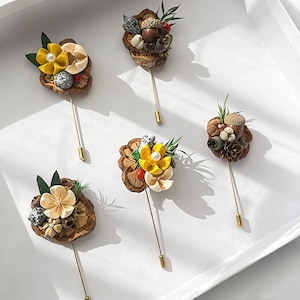 Könnte beinhalten: Fünf dekorative Broschen mit floralen und natürlichen Elementen. Jede Brosche hat eine goldfarbene Nadel und ist mit gelben, cremefarbenen und braunen Blumen sowie kleinen dekorativen Akzenten verziert. Die Broschen sind auf einer weißen Oberfläche angeordnet.