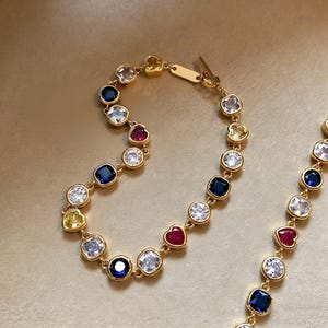 Könnte beinhalten: Ein goldfarbenes Armband mit Knebelverschluss, verziert mit einer Reihe farbiger Edelsteine. Die Edelsteine umfassen klare, blaue, gelbe und rote herzförmige und quadratisch geschliffene Steine. Das Armband wird vor einem neutralen Hintergrund präsentiert.